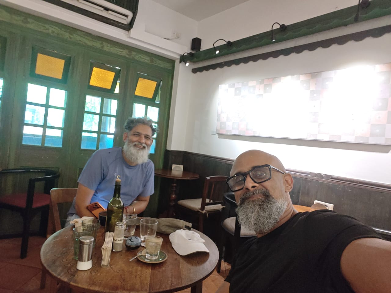 Two men at a wooden table in a Pondicherry café, green shuttered windows behind them, coffee glasses on the table.