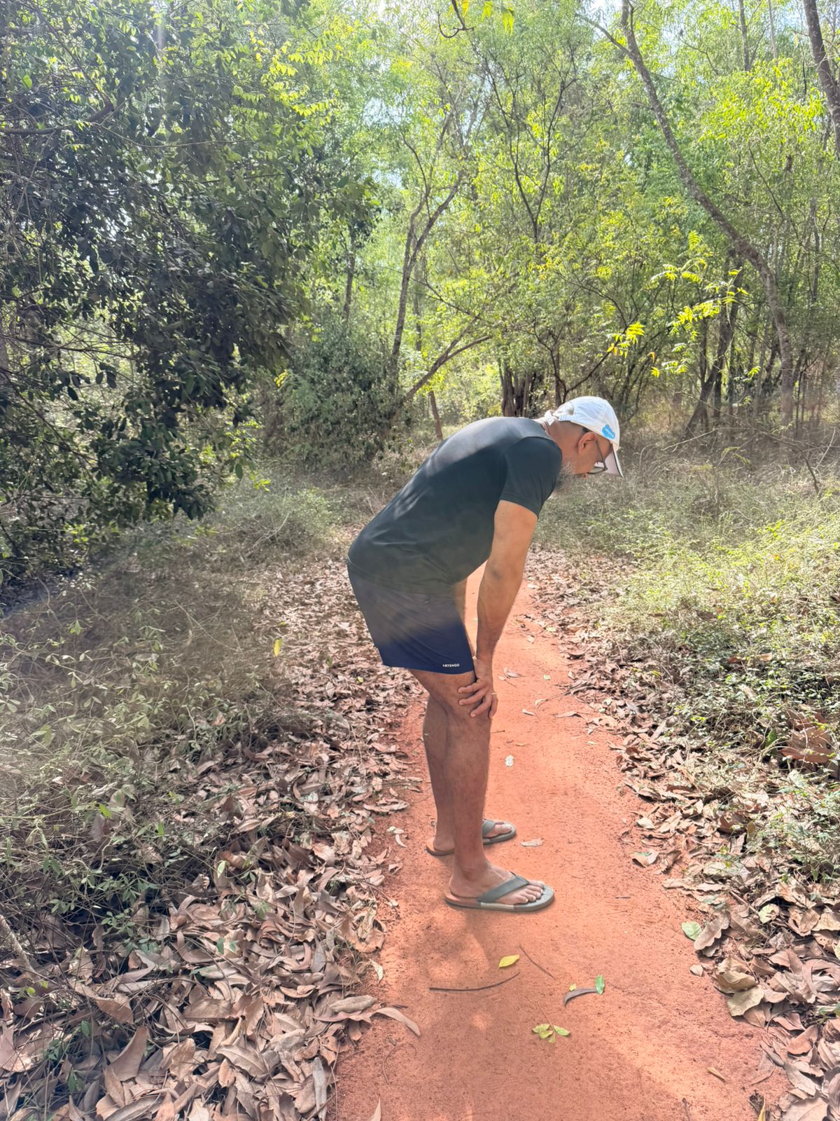 A man bent over, looking closely at something on a red dirt path through green Auroville scrub.